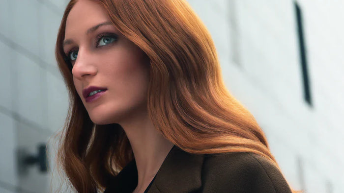 Frau mit langen, glänzenden roten Haaren blickt elegant zur Seite, im Hintergrund eine moderne graue Wand.
