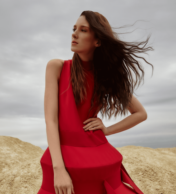 Frau in rotem Kleid mit langem braunem Haar, das vom Wind verweht wird, draußen auf Sand.