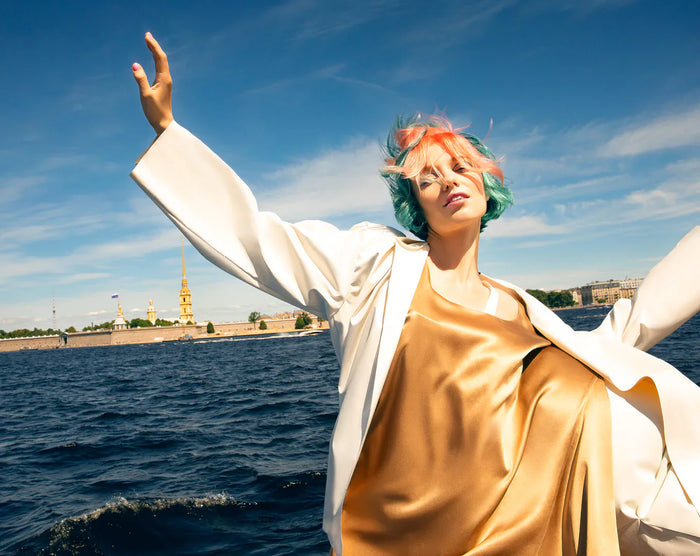 Frau mit kurzem, bunt gefärbtem Haar in Blau und Pink, goldfarbenem Kleid und weißem Mantel posiert am Wasser vor blauem Himmel.