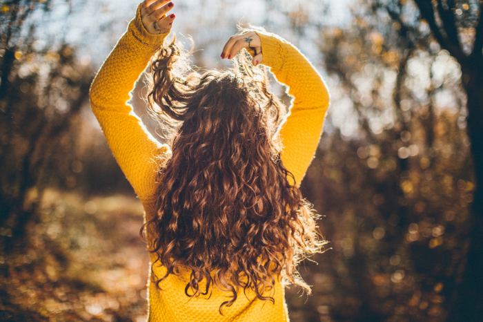 Frau mit langen lockigen Haaren im Sonnenlicht, trägt gelben Pullover und hebt die Arme im herbstlichen Wald.