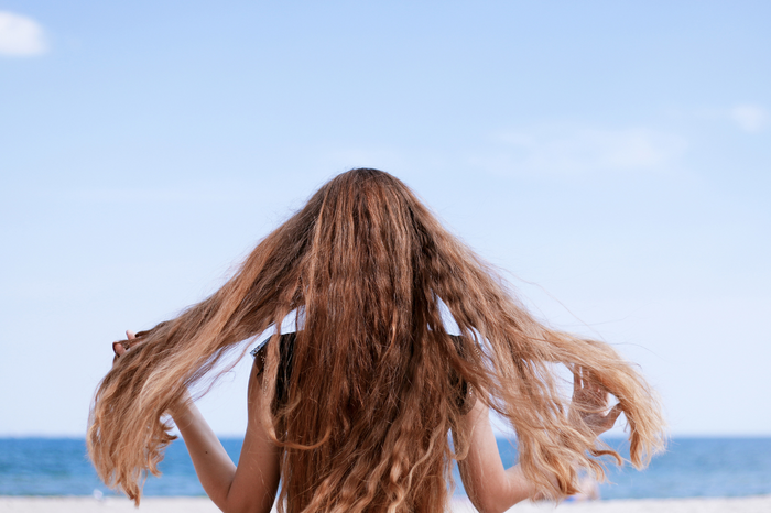 Frau mit langen welligen Haaren am Strand, Rückenansicht zum Meer.