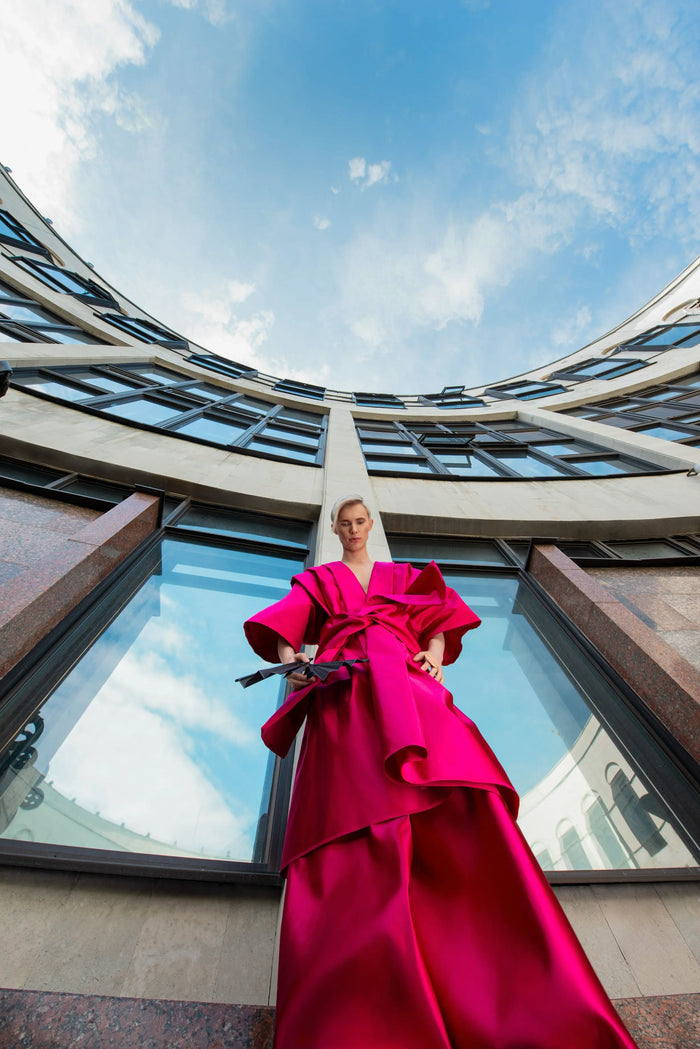 Person in leuchtend rotem Gewand mit schwarzem Fächer posiert vor moderner Architektur, im Hintergrund blauer Himmel mit Wolken.
