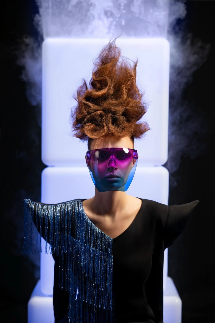 Frau mit avantgardistischer Hochsteckfrisur und pink-blauer Brille vor leuchtenden Würfeln.