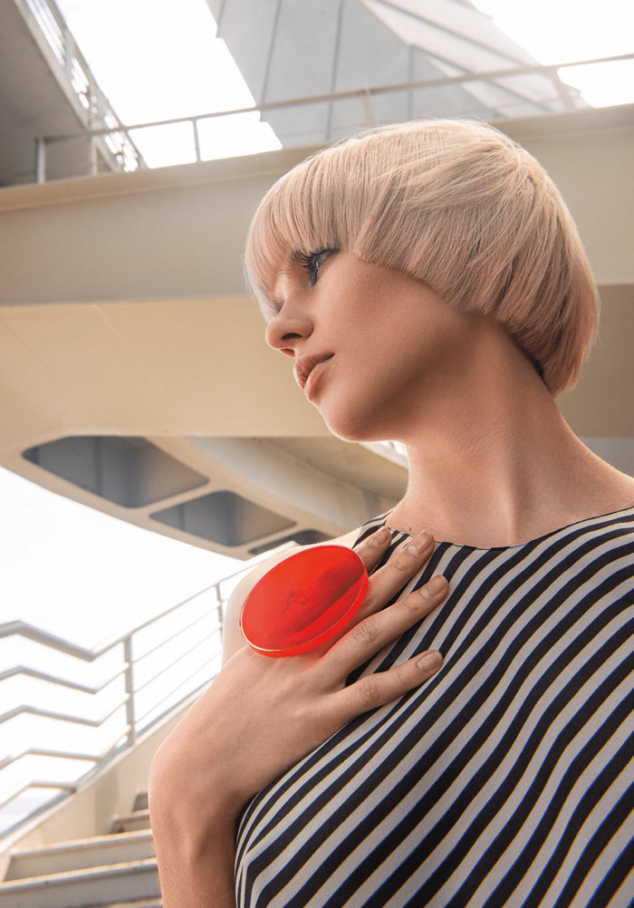 Model mit blondem Bob trägt gestreiftes Kleid und großen roten Ring, posiert vor moderner Treppenkulisse.