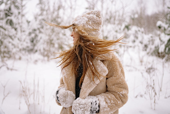 Frau in Winterjacke und Mütze steht im Schnee, während ihr langes Haar vom Wind verweht wird.