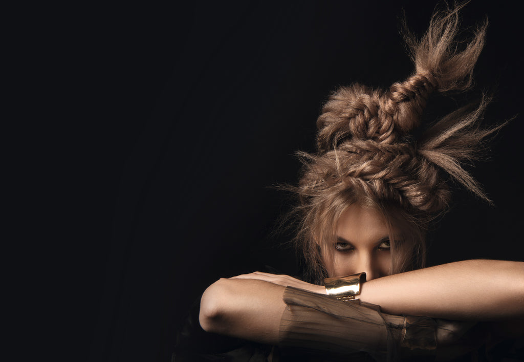 Person mit kunstvoll geflochtener, avantgardistischer Hochsteckfrisur blickt intensiv über ihre verschränkten Arme, dunkler Hintergrund betont das Haarstyling.