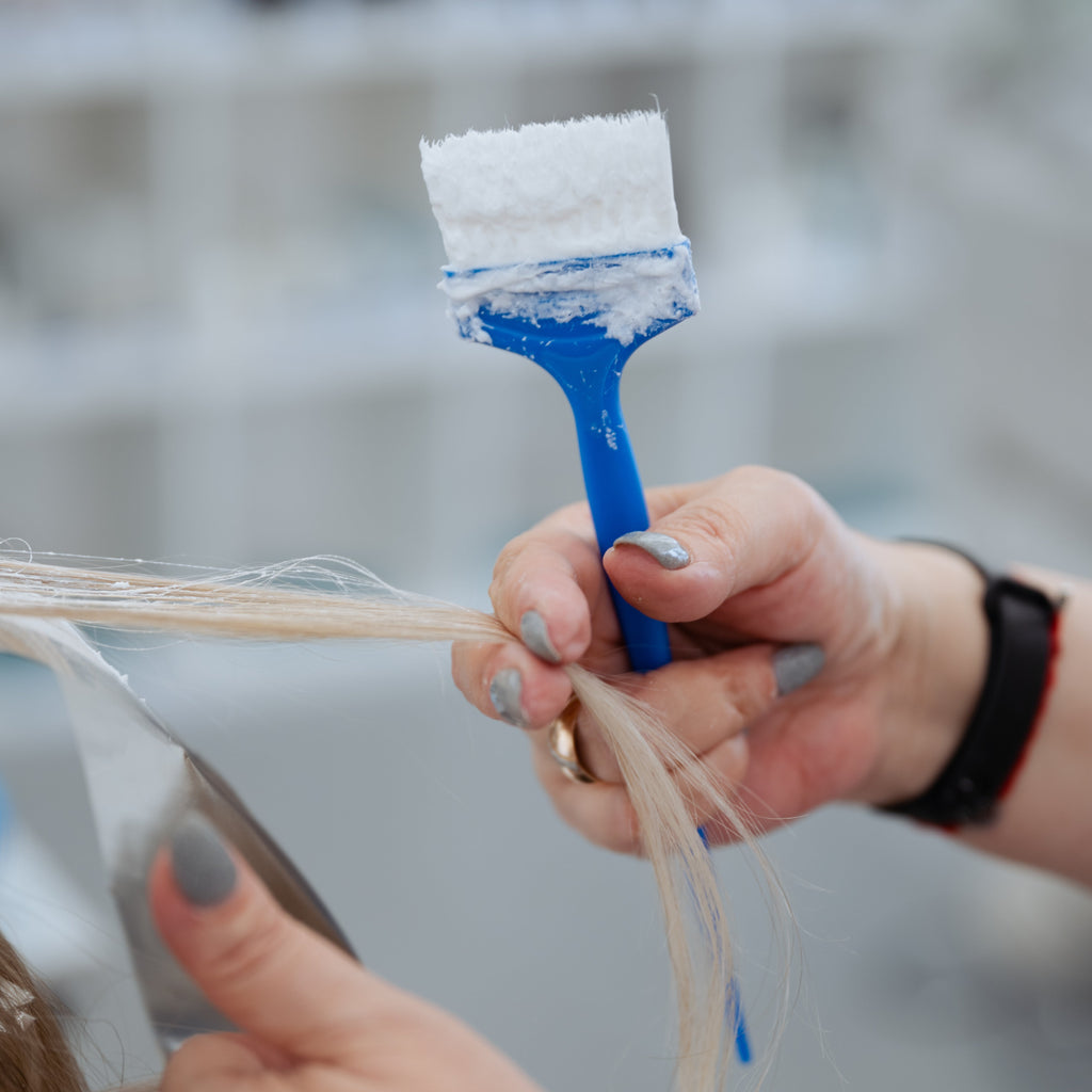 Friseurin trägt mit einem Pinsel Blondierung auf eine Haarsträhne auf, die über eine Folie gelegt wird. Nahaufnahme der Anwendung im Salon.