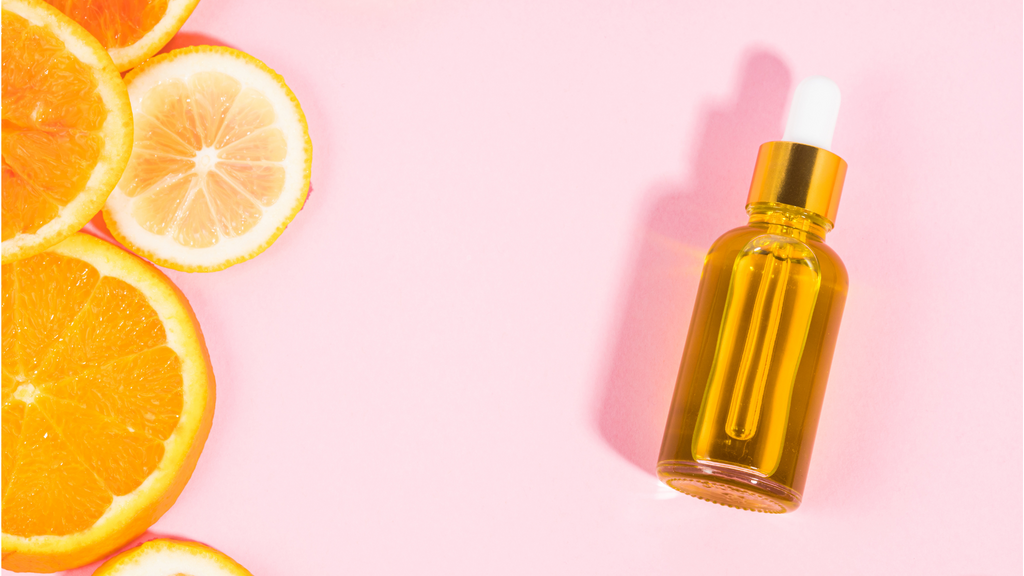 Dekorative Darstellung von Vitamin E: Orangenscheiben und eine Glasflasche mit Pipette, gefüllt mit goldenem Öl, auf rosa Hintergrund.