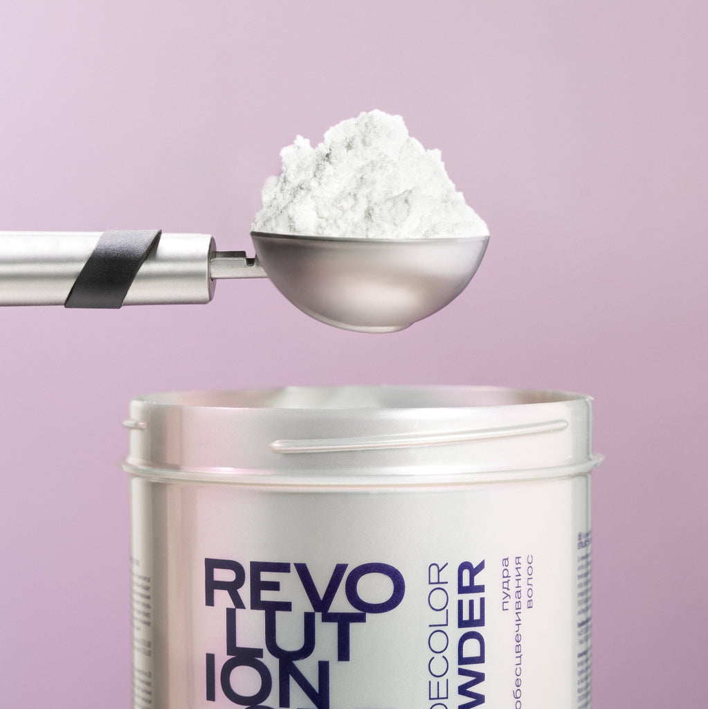 Weiße Dose ESTEL DeLuxe Revolution Blondierpulver, geöffnet mit Metallschaufel voller weißem Pulver, vor rosafarbenem Hintergrund.