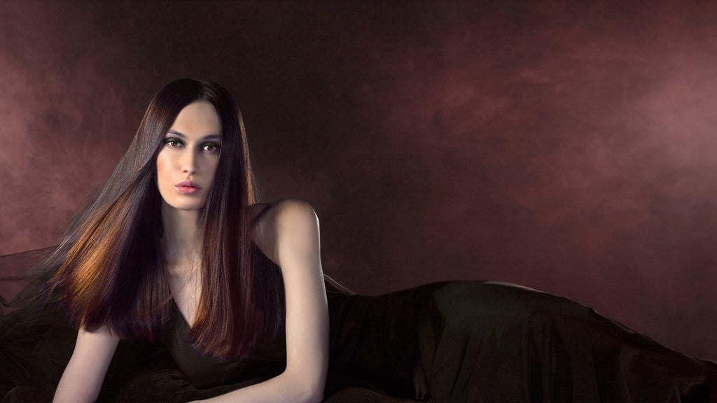 Frau mit sehr langem, glattem dunkelbraunem Haar mit kupferfarbenen Reflexen, in schwarzem Kleid vor dunkelrot schimmerndem Hintergrund.