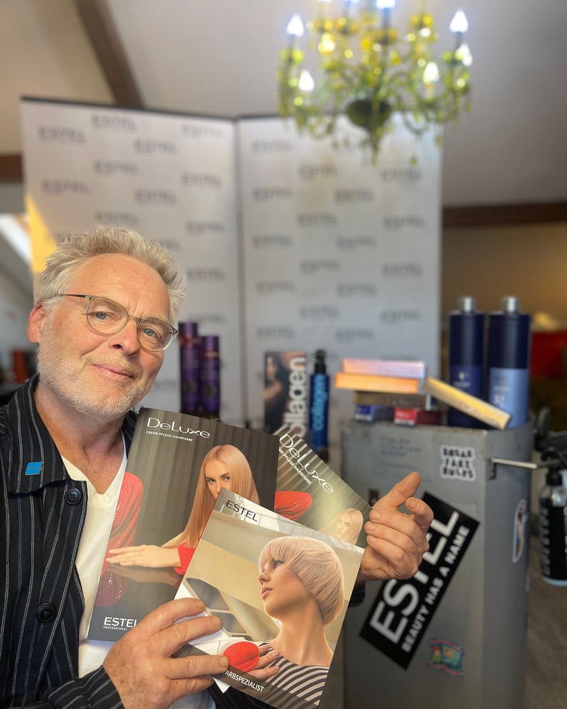 Mann mit Brille hält mehrere ESTEL DeLuxe Farbkarten in den Händen, im Hintergrund ein Messestand mit ESTEL-Logo und Haarpflegeprodukten.