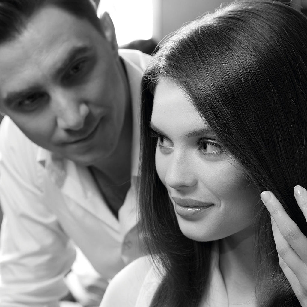 Mann im weißen Hemd betrachtet aufmerksam eine Frau mit glattem, gepflegtem Haar, die sich eine Strähne zwischen den Fingern anschaut.