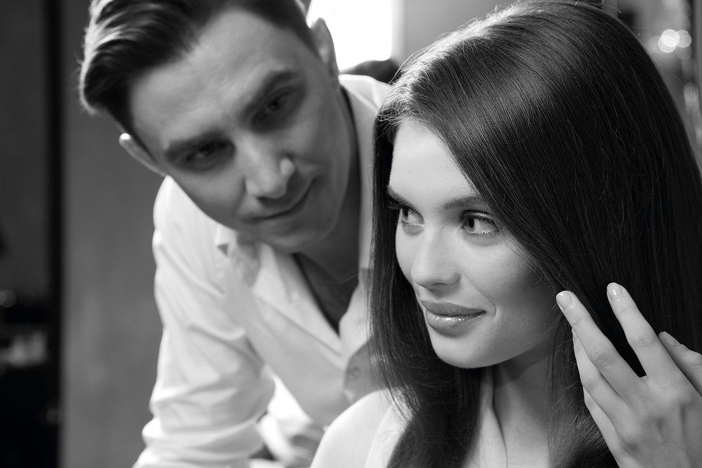 Mann im weißen Hemd betrachtet aufmerksam eine Frau mit glattem, gepflegtem Haar, die sich eine Strähne zwischen den Fingern anschaut.