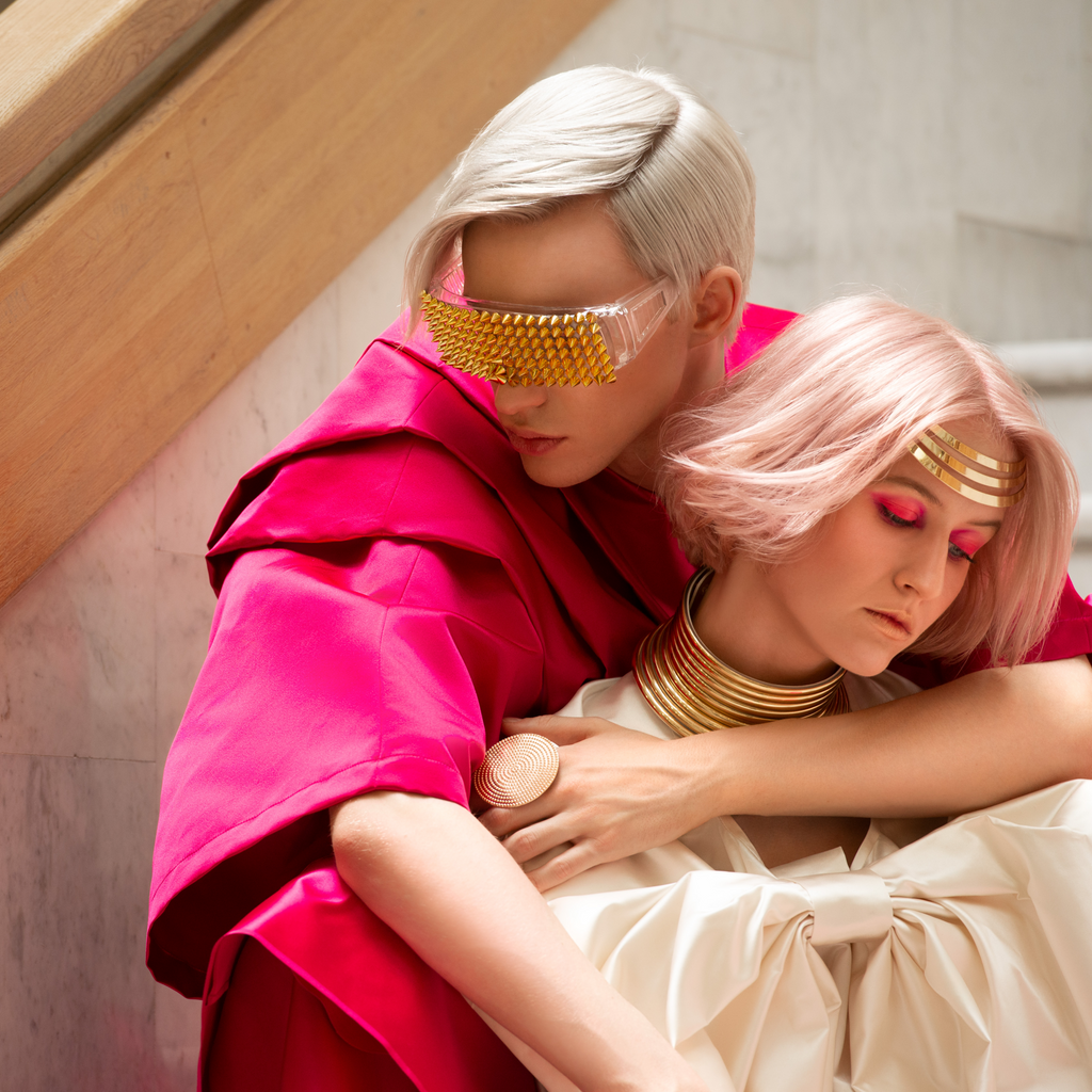 Zwei Personen in futuristischer Mode: eine mit kurzem, silberblondem Haar, golden verzierten Sonnenbrillen und pinkem Oberteil; die andere mit rosafarbenem Bob, goldenen Schmuckelementen und intensiv pinkem Lidschatten. Künstlerisches, avantgardistisches Styling.