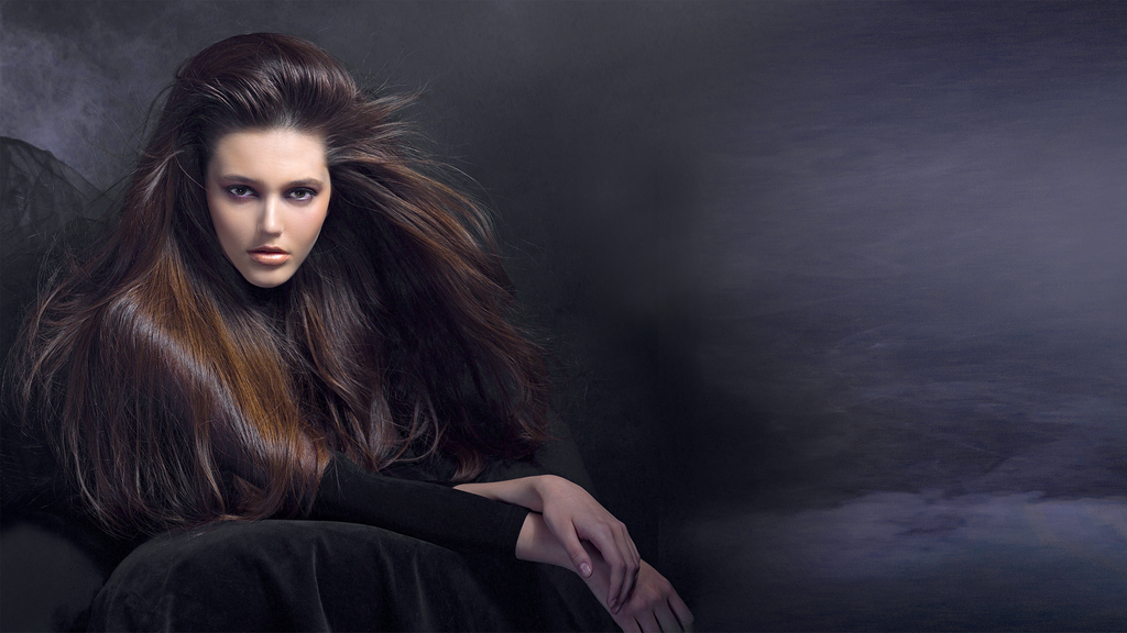 Frau mit sehr langem, voluminösem und glänzendem dunkelbraunem Haar, das über die Schulter fällt, vor dunklem Hintergrund.