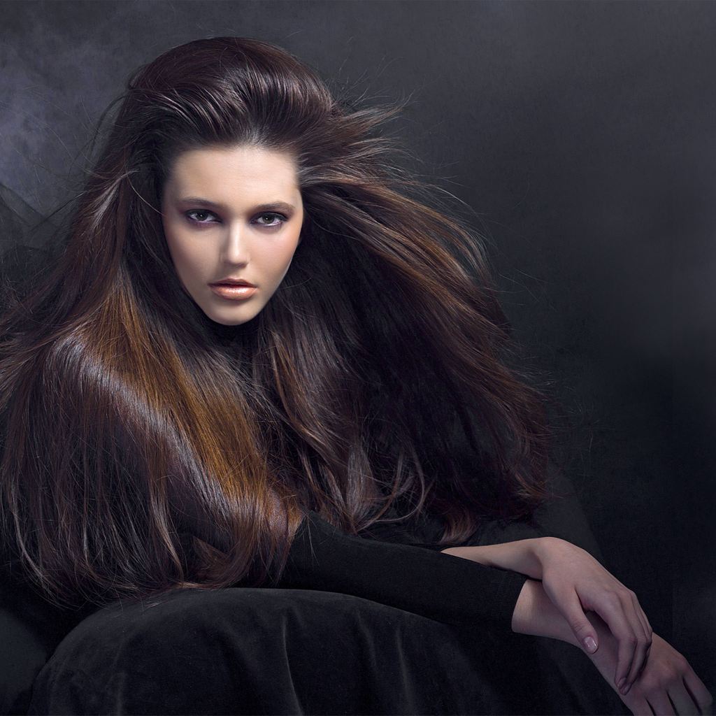 Frau mit sehr langem, voluminösem und glänzendem dunkelbraunem Haar, das über die Schulter fällt, vor dunklem Hintergrund.