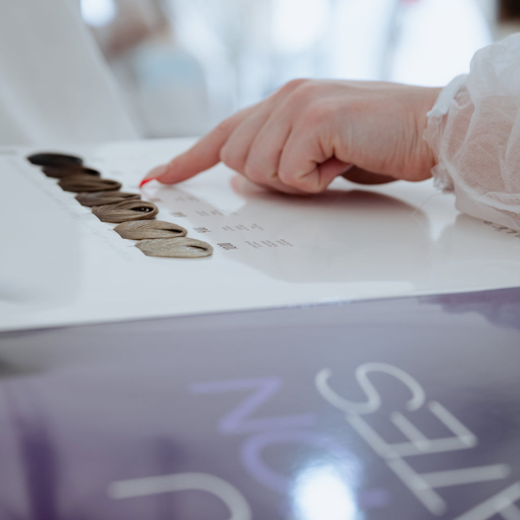 Nahaufnahme einer Hand mit rotem Nagellack, die auf eine Farbkarte mit Haarsträhnen in verschiedenen Blond- und Brauntönen zeigt. Im Vordergrund ist unscharf eine violette Broschüre zu erkennen.