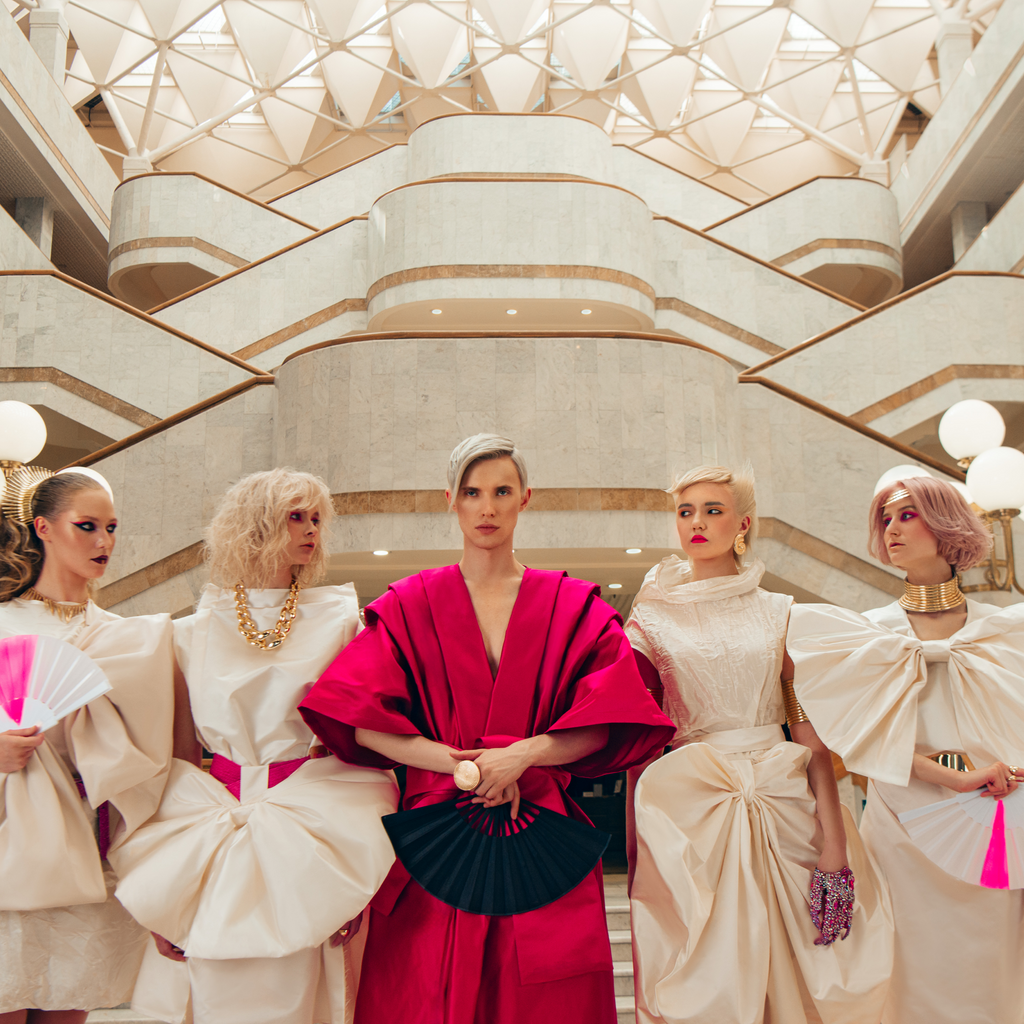 Gruppe von fünf Models in kunstvollen Outfits, eines in kräftigem Pink, die anderen in weißen Kleidern mit großen Drapierungen, posierend vor einer modernen Marmortreppe.
