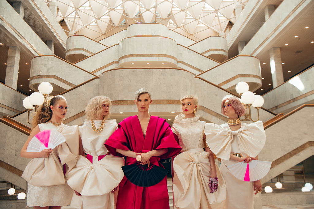 Gruppe von fünf Models in kunstvollen Outfits, eines in kräftigem Pink, die anderen in weißen Kleidern mit großen Drapierungen, posierend vor einer modernen Marmortreppe.