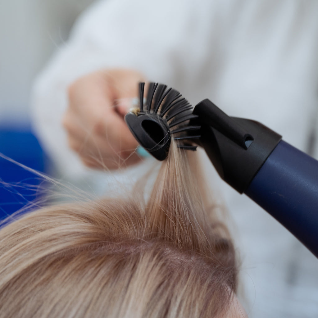 Nahaufnahme einer Friseurin, die mit Föhn und Rundbürste Volumen am Haaransatz einer Kundin föhnt.