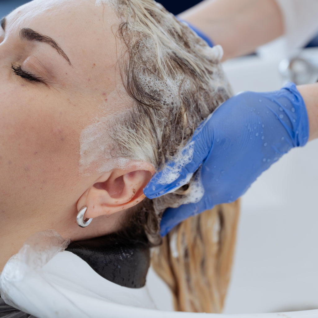 Nahaufnahme einer Frau beim Haarewaschen im Salon, die Kopfhaut wird von einer Friseurin mit blauen Handschuhen und Shampoo sanft massiert.