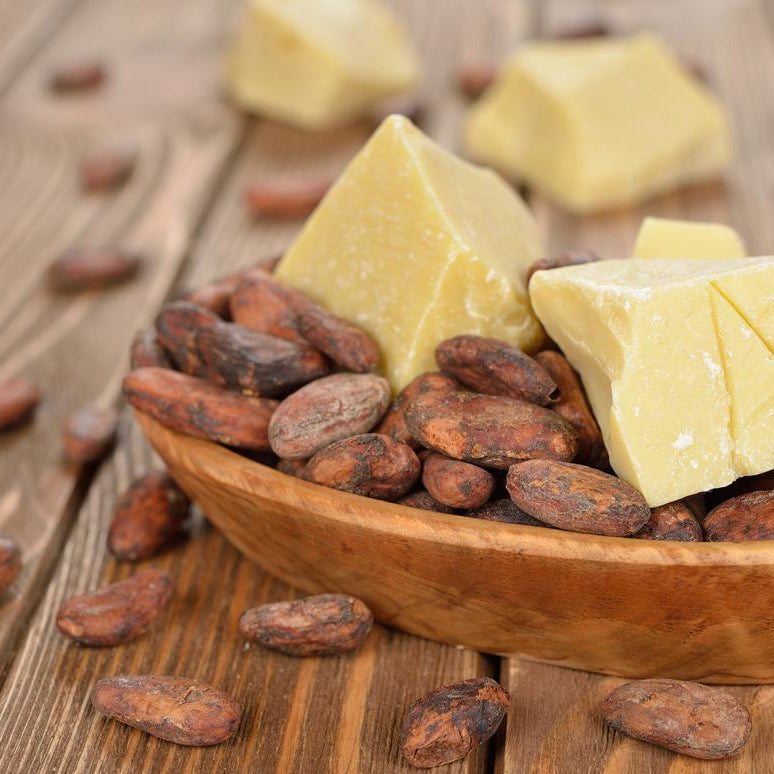 Holzschale mit braunen Kakaobohnen und großen gelblichen Stücken Kakaobutter, arrangiert auf einem Holztisch