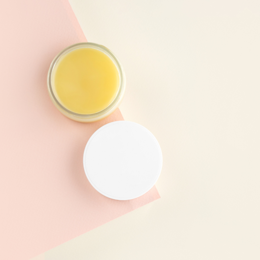 Dekoratives Glasgefäß mit gelblichem Lanolin, daneben ein weißer Deckel auf pastellfarbenem Hintergrund