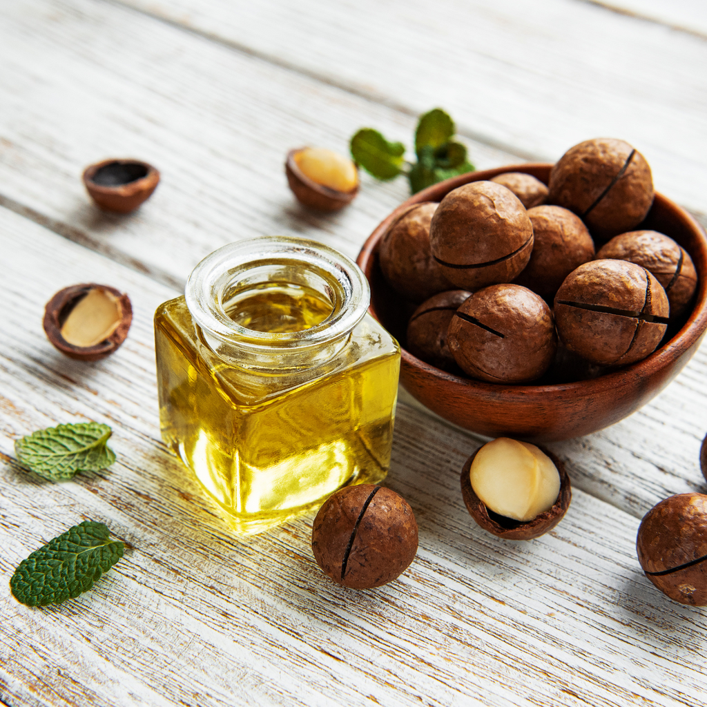 Kleines quadratisches Glasfläschchen mit goldenem Öl neben einer Holzschale voller Macadamianüsse, auf weißem Holztisch mit Minzblättern dekoriert.
