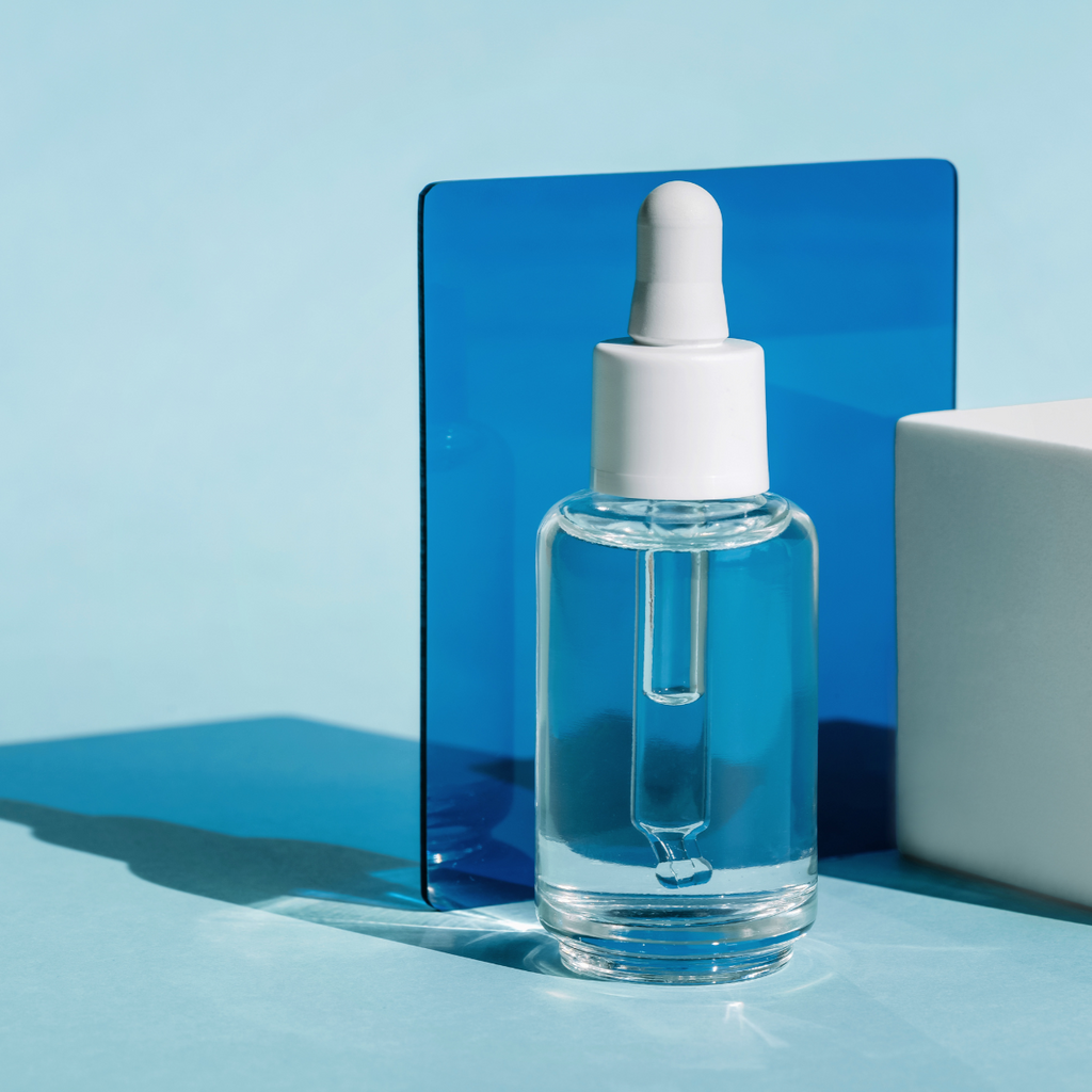 Transparente Glasflasche mit weißer Pipette, gefüllt mit hellblauer Flüssigkeit, vor hellem Hintergrund mit blauer Platte und weißem Block.