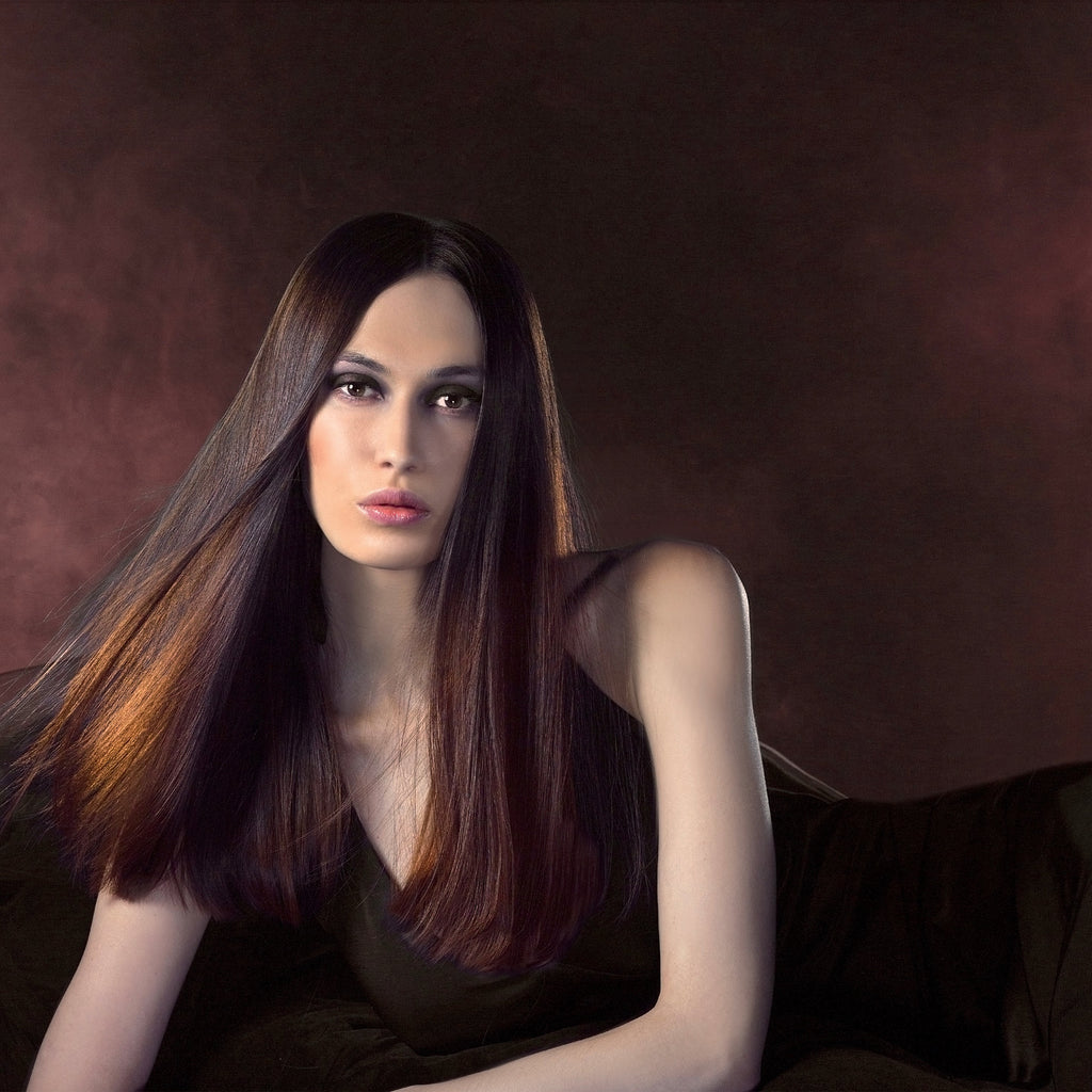 Frau mit sehr langem, glattem dunkelbraunem Haar mit kupferfarbenen Reflexen, in schwarzem Kleid vor dunkelrot schimmerndem Hintergrund.