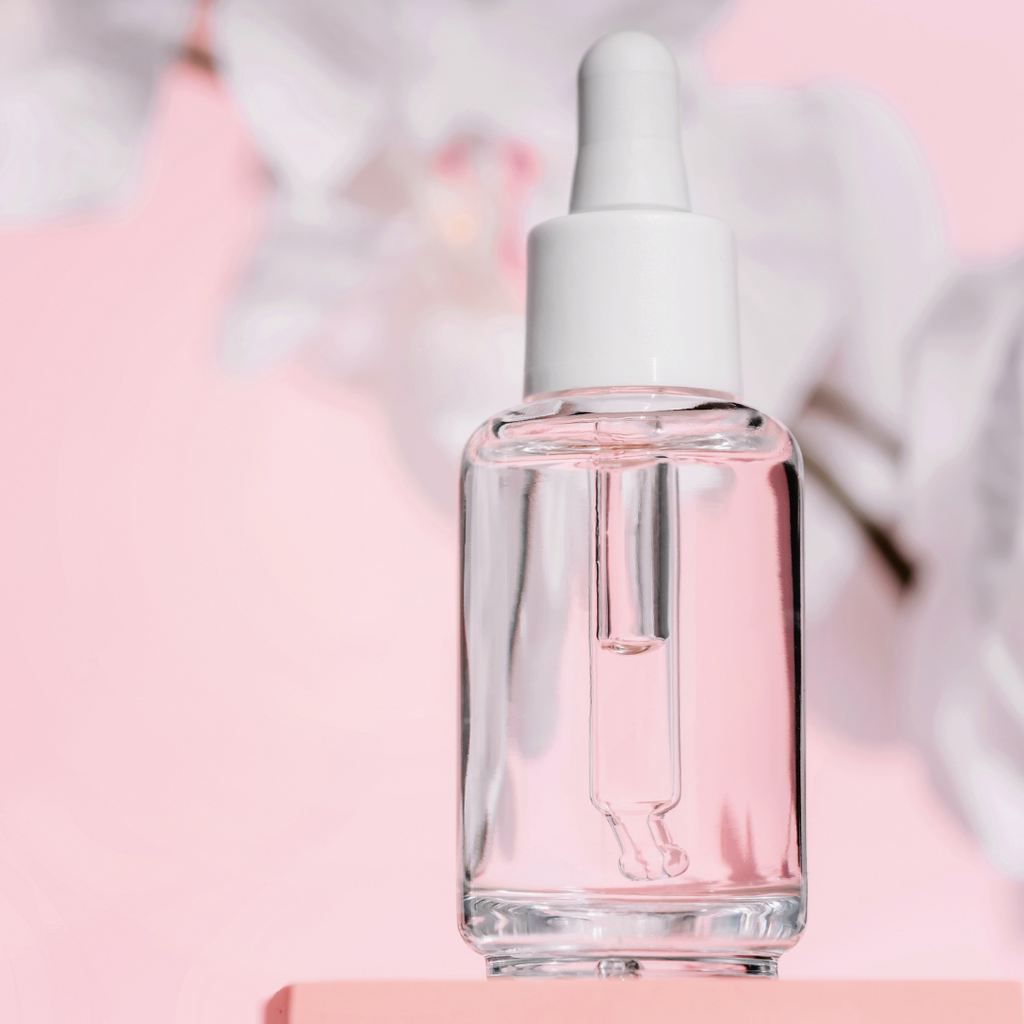 Transparente Glasflasche mit weißer Pipette, gefüllt mit roséfarbener Flüssigkeit, vor rosa Hintergrund mit unscharfen Blüten.