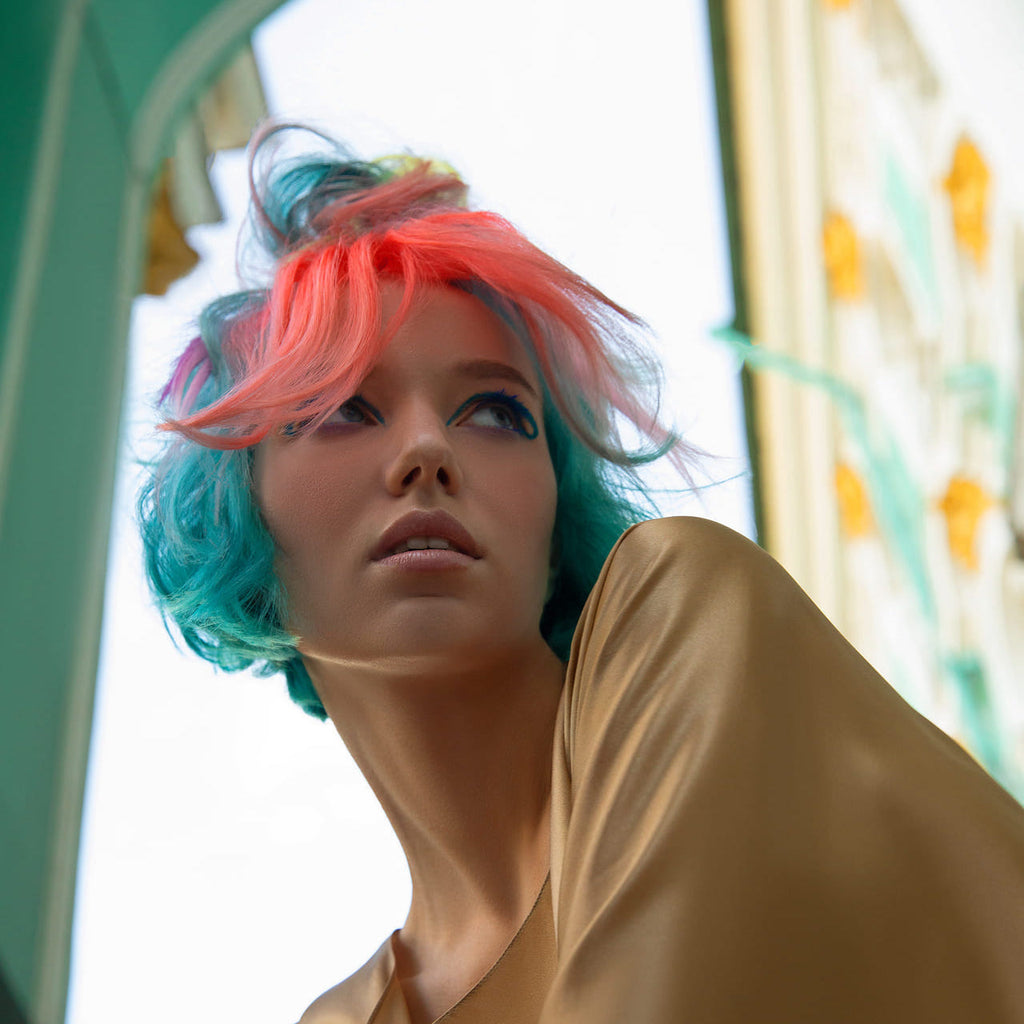 Nahaufnahme einer Frau mit türkis-blauen Haaren und pinken Strähnen, goldener Kleidung und auffälligem Eyeliner vor architektonischem Hintergrund