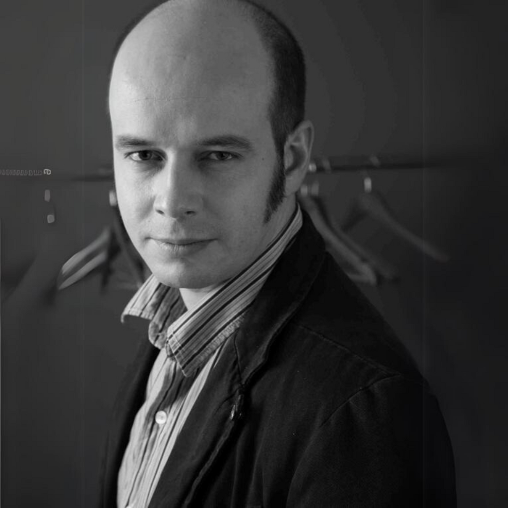 Schwarz-weiß Porträt von Lev Okhotin im Sakko und Hemd, im Hintergrund Kleiderbügel an einer Garderobe