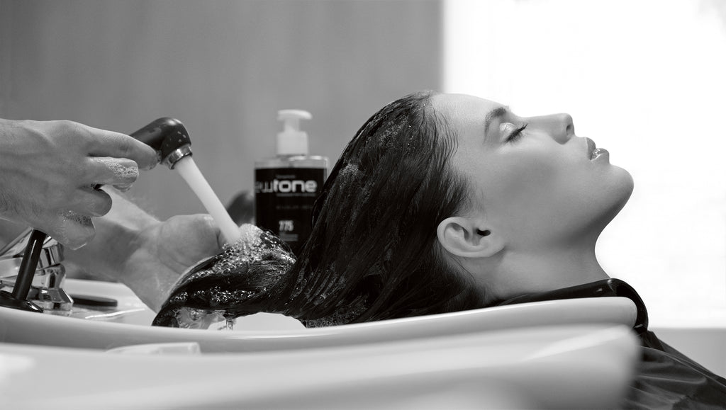 Frau lehnt entspannt am Waschbecken im Salon, während ein Friseur ihre Haare mit Wasser auswäscht – professionelle Haarbehandlung im Einsatz.