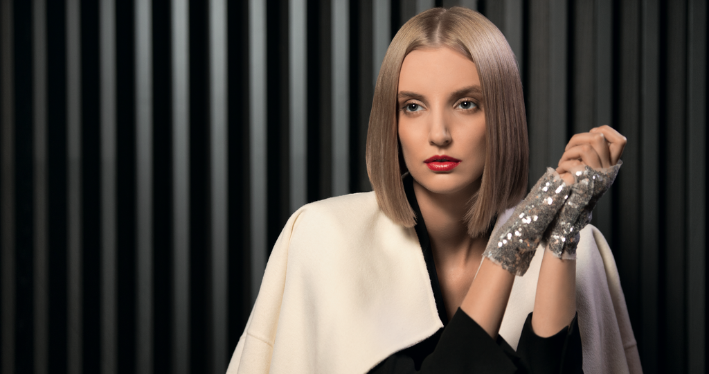 Frau mit glattem blondem Bob und rotem Lippenstift trägt weiße Jacke und glitzernde Handschuhe vor dunklem Hintergrund
