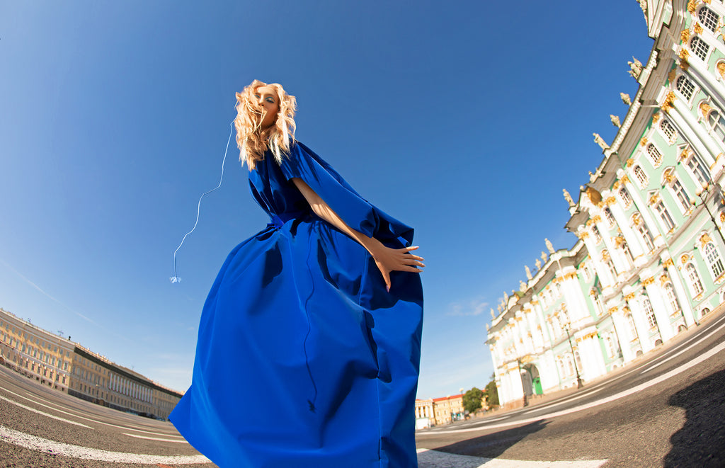 Frau mit langen blonden Haaren in weitem blauen Kleid posiert auf dem Palastplatz vor dem Winterpalast unter blauem Himmel