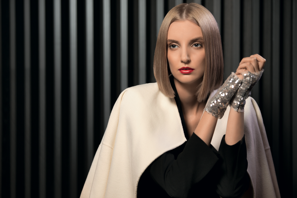 Frau mit glattem blondem Bob und rotem Lippenstift trägt weiße Jacke und glitzernde Handschuhe vor dunklem Hintergrund