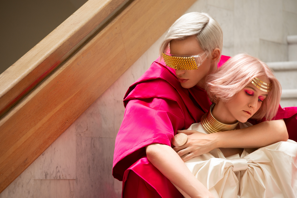 Zwei Personen in futuristischer Mode: eine mit kurzem, silberblondem Haar, golden verzierten Sonnenbrillen und pinkem Oberteil; die andere mit rosafarbenem Bob, goldenen Schmuckelementen und intensiv pinkem Lidschatten. Künstlerisches, avantgardistisches Styling.